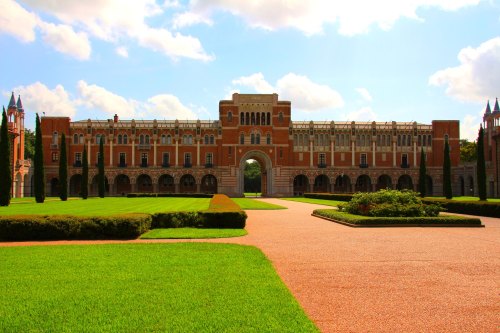 A grand brick building with an archway, surrounded by manicured lawns and trees under a bright blue sky.