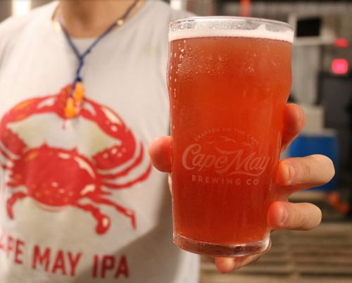 A person holding a pint glass of red beer with a logo from Cape May Brewing Co., wearing a crab-themed shirt.