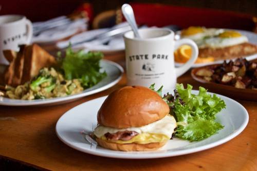 A close-up of a burger with lettuce on a plate, surrounded by various dishes and a coffee mug in a cozy setting.
