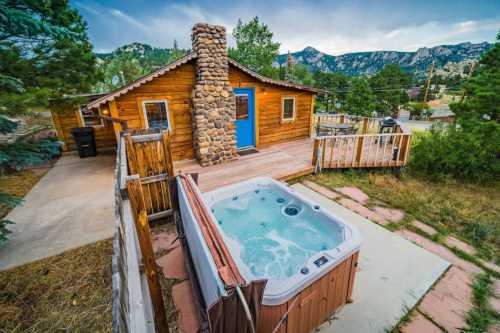 Cozy wooden cabin with a stone chimney, hot tub, and scenic mountain views in a lush outdoor setting.
