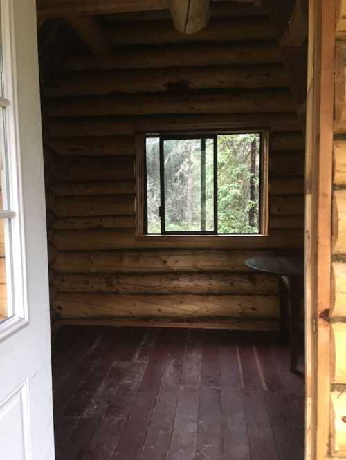 Interior of a rustic log cabin with a window overlooking a forest, featuring wooden walls and a simple table.