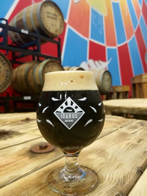 A glass of dark beer with a frothy head sits on a wooden table, with colorful barrels and a mural in the background.