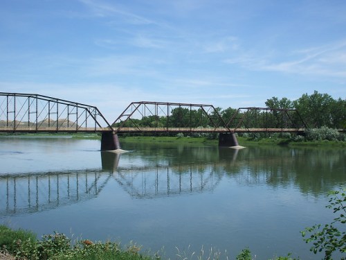 A metal truss bridge spans a calm river, reflecting in the water, surrounded by green trees and a clear blue sky.