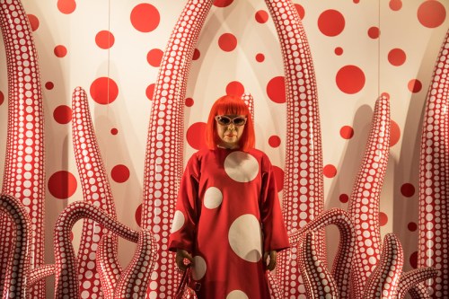 A person in a red polka dot outfit stands against a backdrop of red tentacles and polka dots.