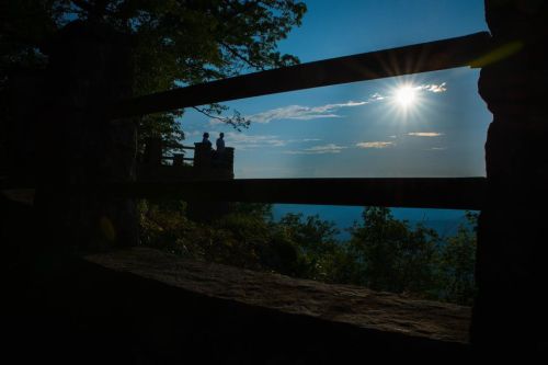 Silhouetted figures stand by a railing, overlooking a scenic view with a bright sun shining in the sky.
