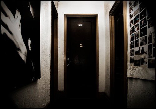 A dimly lit hallway with a black door labeled "211" and a wall covered in photographs.