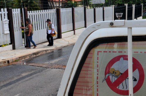 A woman approaches a gate while a man walks by, with a "no mosquitoes" sign visible on a vehicle window.