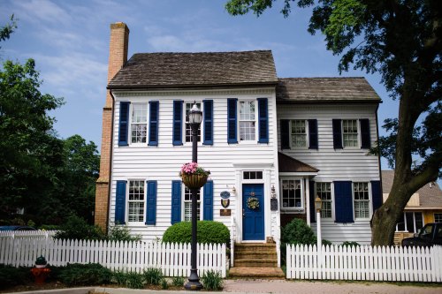 A charming two-story white house with blue shutters, a front porch, and a picket fence, surrounded by greenery.