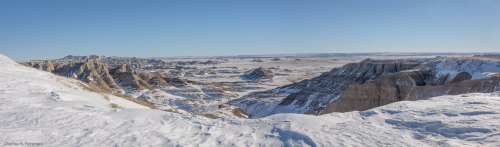 A panoramic view of snow-covered Badlands, showcasing rugged cliffs and vast plains under a clear blue sky.