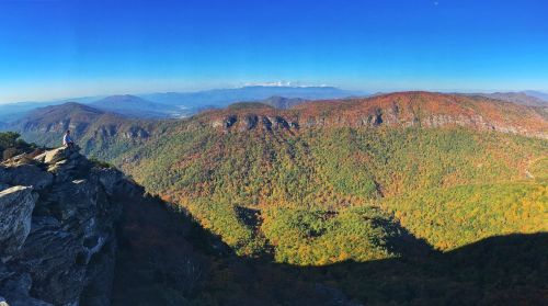 A panoramic view of colorful autumn mountains with a person sitting on a rocky ledge, under a clear blue sky.