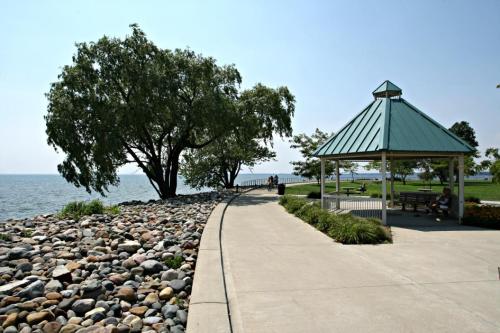 A scenic waterfront path with a gazebo, trees, and a rocky shoreline under a clear blue sky.