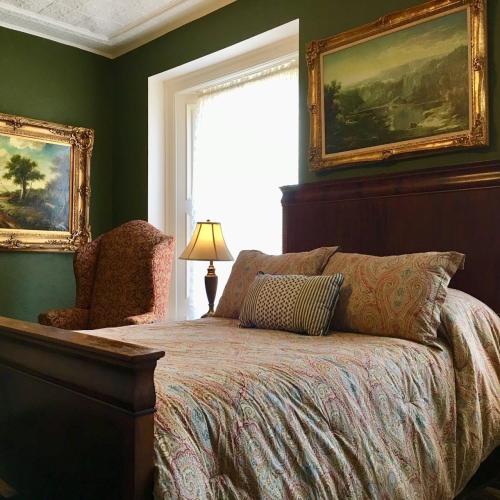 Cozy bedroom with a patterned bedspread, a vintage armchair, and framed landscape paintings on green walls.