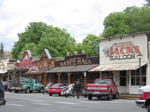 Historic western-style buildings line a street, featuring a dance hall and a saloon, with parked vehicles in front.