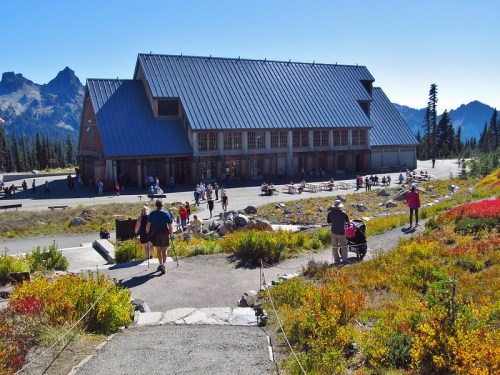 A large building surrounded by colorful foliage and visitors, set against a backdrop of mountains and clear blue sky.