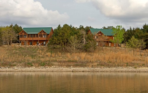 Two wooden cabins with green roofs sit on a grassy hillside by a calm lake, surrounded by trees and cloudy skies.