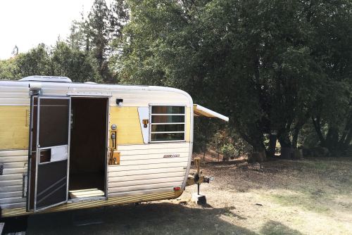 A vintage silver trailer parked under trees, with an open door and a sunny outdoor setting.