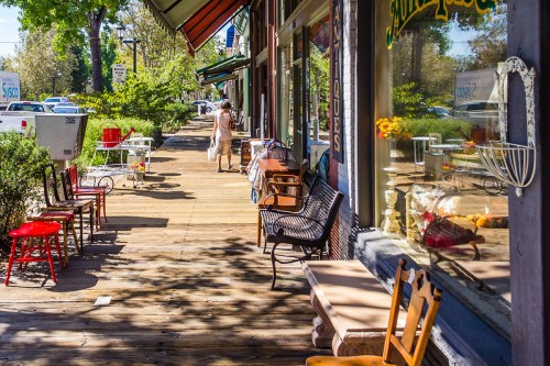 A sunny sidewalk lined with colorful chairs and storefronts, with a person walking in the distance.