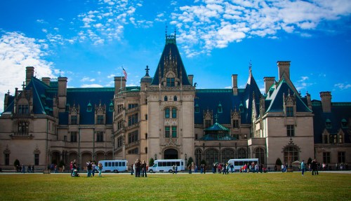 A grand mansion with intricate architecture, surrounded by green grass and visitors, under a bright blue sky.