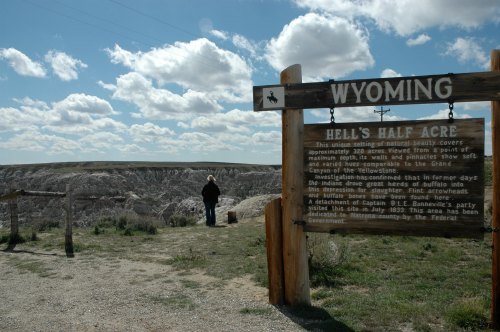 A person stands near a sign for Hell's Half Acre in Wyoming, overlooking a vast, rugged landscape under a cloudy sky.