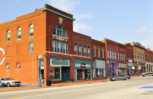 Historic brick buildings line a street, featuring shops and a music hall, with American flags displayed.