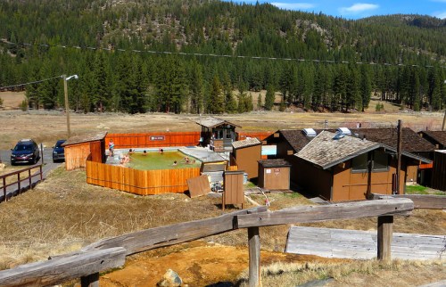 A scenic view of a rustic lodge with a small pool, surrounded by wooden fences and lush green trees in the background.