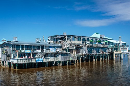 A wooden pier with shops and restaurants overlooking a calm, brown river under a clear blue sky.