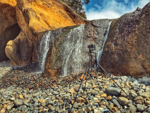 A camera on a tripod near a waterfall, surrounded by rocky terrain and cliffs under a blue sky.
