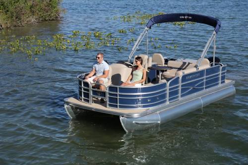 A couple sits on a pontoon boat, fishing on a calm lake surrounded by greenery.
