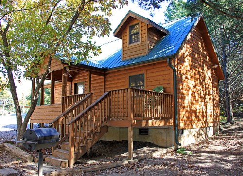 A cozy wooden cabin with a green metal roof, surrounded by trees and featuring a porch and grill.