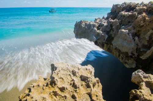 Waves gently wash over rocky shore with a boat in the distance under a clear blue sky.