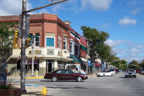 A street view of a small town with shops, cars, and trees under a partly cloudy sky.