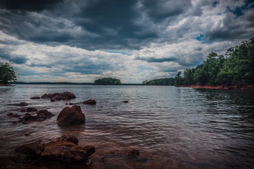 A serene lake scene with rocky shorelines, dark clouds overhead, and lush greenery in the background.