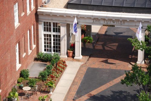 Entrance of The Tutwiler hotel, featuring brick pathways, potted plants, and flags, surrounded by greenery.