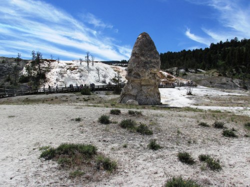 A large rock formation stands in a sandy area with sparse vegetation, under a blue sky with wispy clouds.