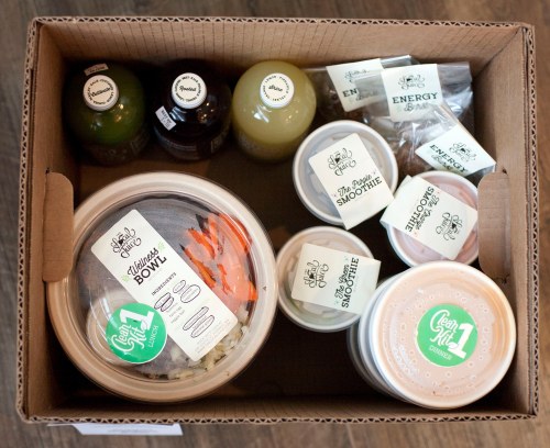 A cardboard box containing healthy meal options, including bowls, smoothies, and drinks, all labeled with clean eating tags.