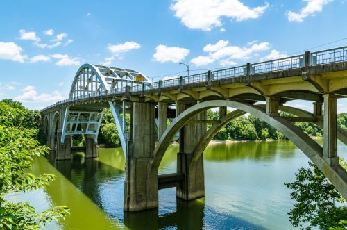 A modern arch bridge spans a river, reflecting blue skies and clouds, surrounded by lush greenery.