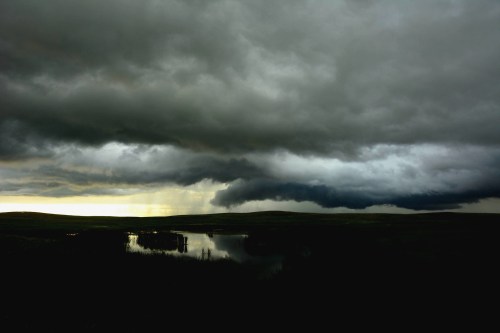 Dark, dramatic clouds loom over a calm lake, reflecting the moody sky and surrounding landscape.
