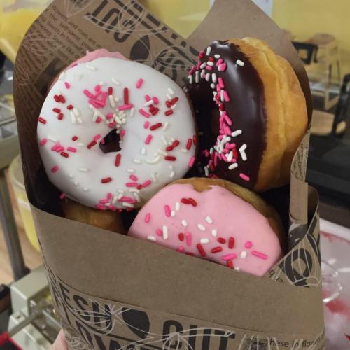 A paper bag filled with four colorful donuts, including chocolate and pink frosted varieties, topped with sprinkles.