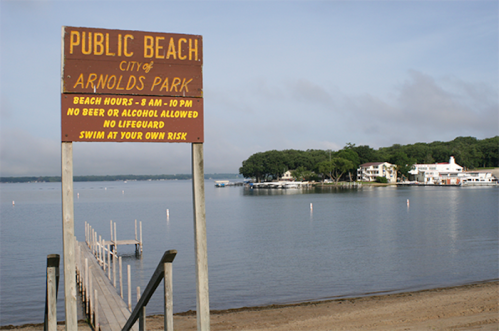 Sign for Public Beach at Arnolds Park, with a wooden dock leading to a calm lake and houses in the background.