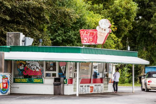 A small ice cream shop with a green roof and a neon sign, featuring a customer at the window on a cloudy day.