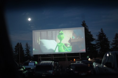 A drive-in movie screen shows a green character at night, with a full moon and parked cars in the foreground.