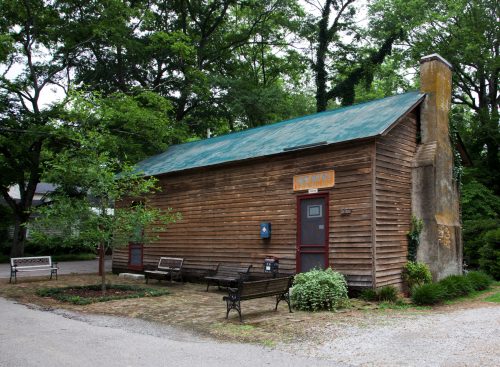 A rustic wooden building with a green roof, surrounded by trees and benches, in a peaceful outdoor setting.