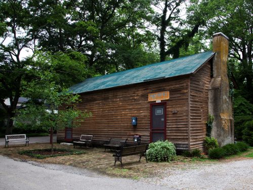 A rustic wooden building with a green roof, surrounded by trees and benches, in a peaceful outdoor setting.