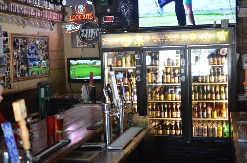 A bar interior featuring a beer fridge, taps, and TVs displaying sports, with a cozy atmosphere and wooden decor.