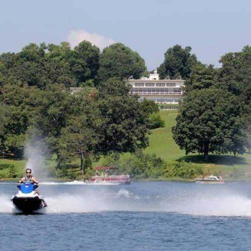 A person rides a jet ski on a lake, with a large house and trees in the background on a sunny day.