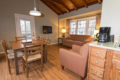 Cozy living space with a dining table, sofa, TV, and kitchenette, featuring wooden beams and large windows.