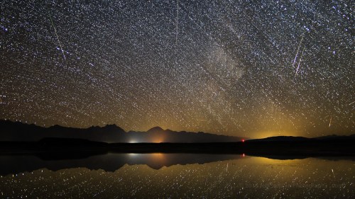 A starry night sky filled with meteors, reflecting on a calm lake with mountains in the background.