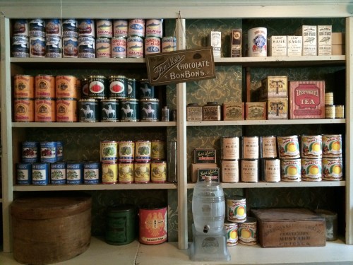 A vintage display of assorted canned goods and packaged items on wooden shelves, featuring colorful labels and a glass water dispenser.
