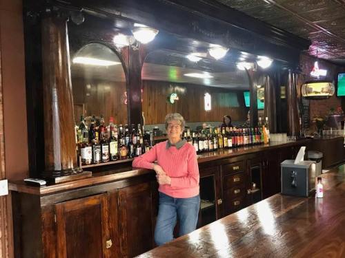 A woman in a pink sweater stands at a wooden bar, smiling beside a variety of liquor bottles on the shelves.
