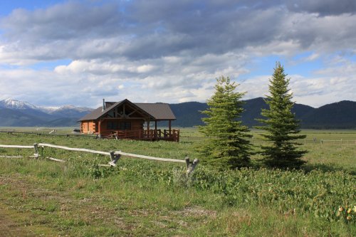 A rustic cabin surrounded by green fields and two evergreen trees, with mountains and cloudy skies in the background.